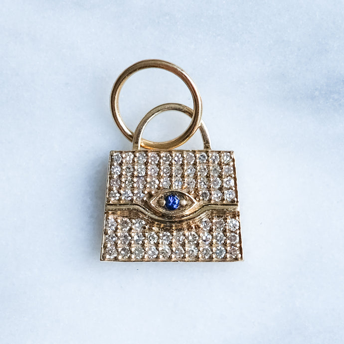 Gold purse shaped pendant with diamond-encrusted design and blue gemstone on a light blue background